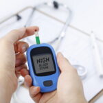 Hand holding a blood glucose meter measuring blood sugar, the background is a stethoscope and chart file